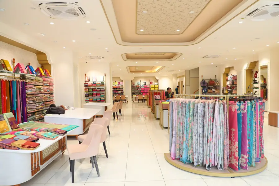 The ground floor: white tiled floor, cove-lit ceiling, rails of printed dress material and seating for customers.